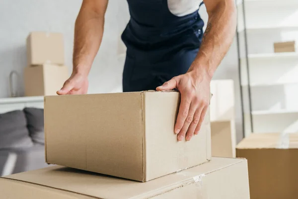 Partial view of tattooed mover in overalls holding box in apartment — Stock Photo