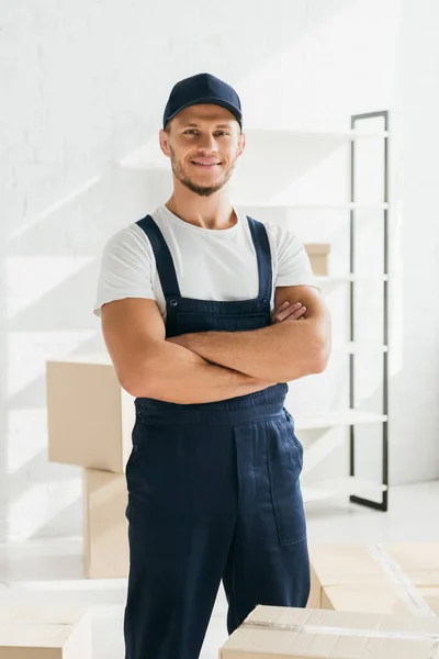 Happy mover in uniform and cap standing with crossed arms in apartment — Stock Photo
