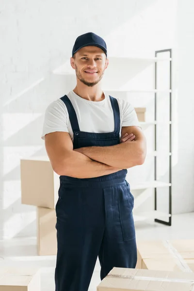 Felice mover in uniforme e cappuccio in piedi con le braccia incrociate in appartamento — Foto stock