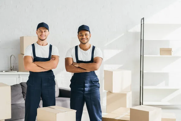Веселые грузчики в шапках, стоящие со скрещенными руками возле коробок в комнате — стоковое фото