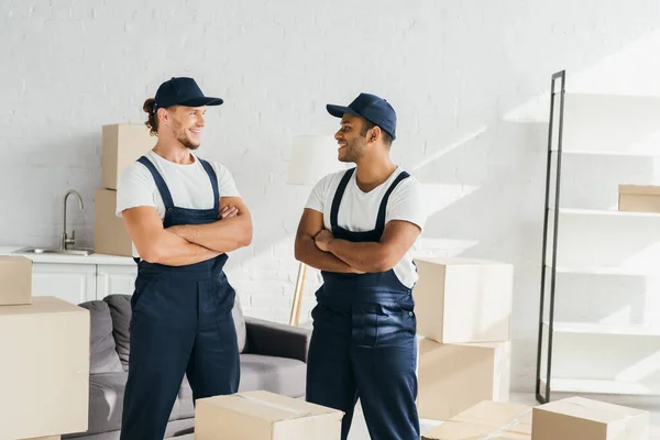 Fröhliche Umzugsteilnehmer in Mützen, die mit verschränkten Armen stehen, während sie einander in der Nähe von Schachteln im Raum anschauen — Stockfoto