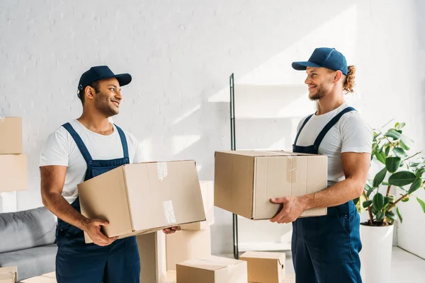 Heureux déménageurs multiethniques dans des casquettes et des boîtes de transport uniformes dans l'appartement — Photo de stock