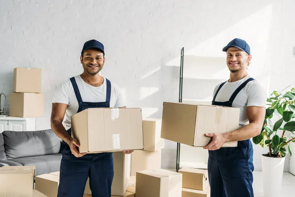 Cheerful multiethnic movers in caps and uniform carrying boxes in apartment — Stock Photo