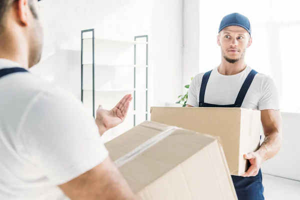 Überraschter Umzugshelfer in Mütze und Uniform, während er einen indischen Kollegen im verschwommenen Vordergrund betrachtet — Stockfoto