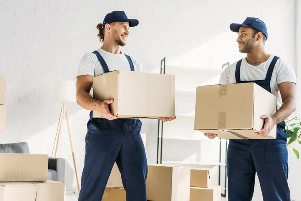 Sorridenti traslocatori multietnici in berretti e scatole di trasporto uniformi in appartamento — Foto stock