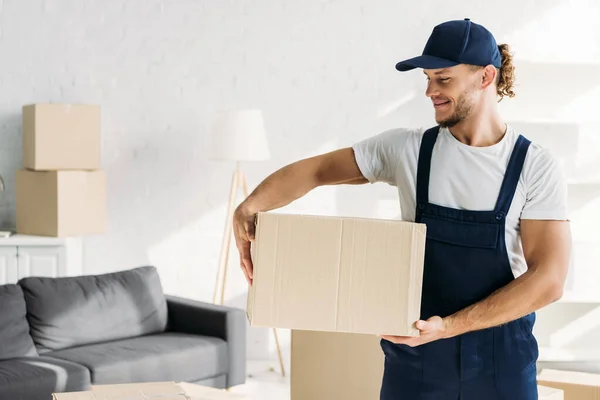 Positive mover in cap and uniform holding carton box in apartment — Stock Photo