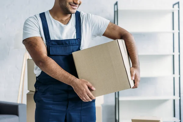 Cropped view of cheerful indian courier in uniform holding box in apartment — Stock Photo