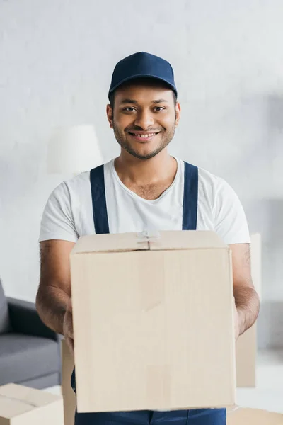 Mensajero indio positivo en gorra y caja de espera uniforme en apartamento - foto de stock