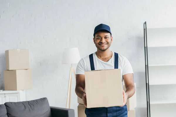 Young cheerful indian courier in cap and uniform holding box in apartment — Stock Photo