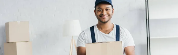 Cheerful indian courier in cap and uniform holding box in apartment, banner — Stock Photo