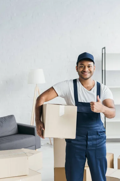 Cheerful indian courier in cap and uniform holding box and showing thumb up in apartment — Stock Photo