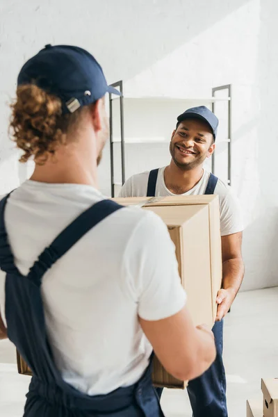 Smiling indian mover in uniform and cap carrying huge carton box with colleague on blurred foreground — Stock Photo