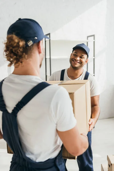 Sorridente mover indiano in uniforme e cappuccio che trasporta enorme scatola di cartone con collega in primo piano sfocato — Foto stock