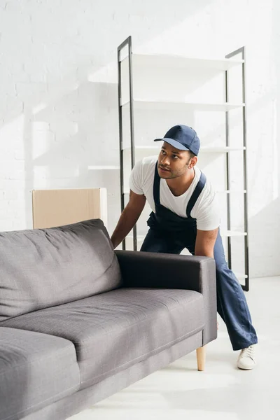 Young indian mover in uniform and cap carrying sofa in apartment — Stock Photo