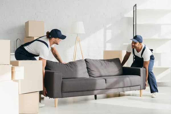 Feliz multicultural movers en uniforme sonriendo mientras llevar sofá en apartamento - foto de stock