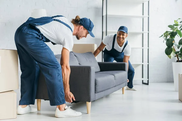 Happy multiethnic movers in uniform smiling while carrying couch in apartment — Stock Photo