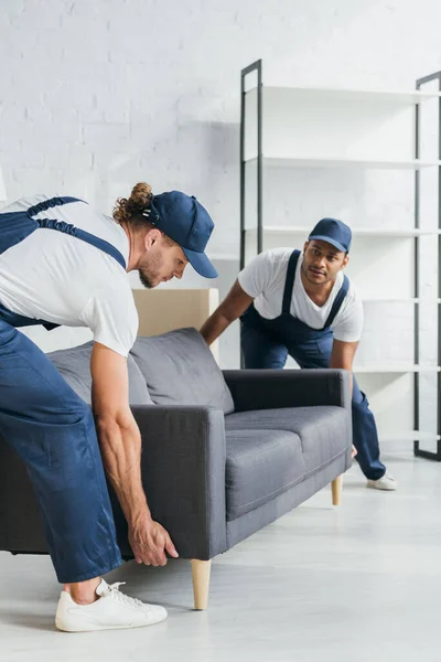 Young multiethnic movers in uniform carrying couch in apartment — Stock Photo