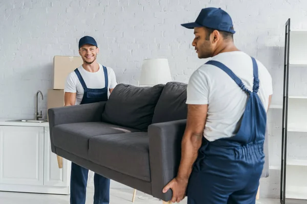 Young mover in uniform smiling while carrying couch with indian colleague on blurred foreground — Stock Photo