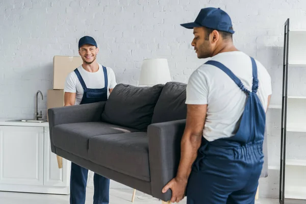 Jovem mover em uniforme sorrindo enquanto carrega sofá com colega indiano em primeiro plano desfocado — Fotografia de Stock