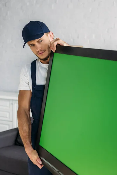 Young mover in uniform carrying plasma tv with green screen in apartment — Stock Photo