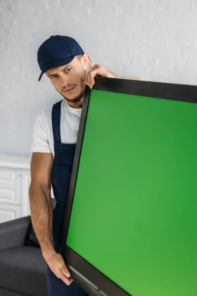 Jeune déménageur en uniforme portant la télévision à écran plasma avec écran vert dans l'appartement — Photo de stock