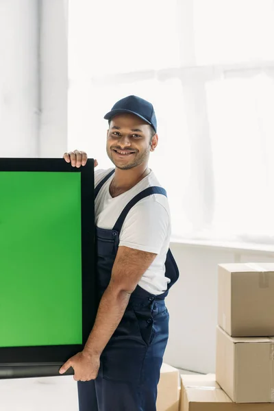 Happy indian mover in uniform carrying plasma tv with green screen in apartment — Stock Photo