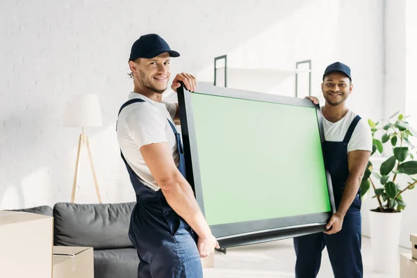 Happy multicultural movers in uniform carrying plasma tv with green screen in apartment — Stock Photo