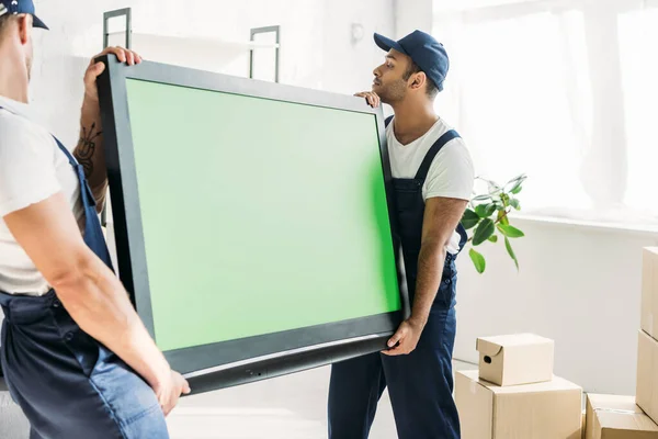 Traslocatori multiculturali in berretti e televisore al plasma con schermo verde in appartamento — Foto stock