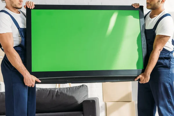 Cropped view of multicultural movers in uniform carrying plasma tv with green screen in apartment — Stock Photo