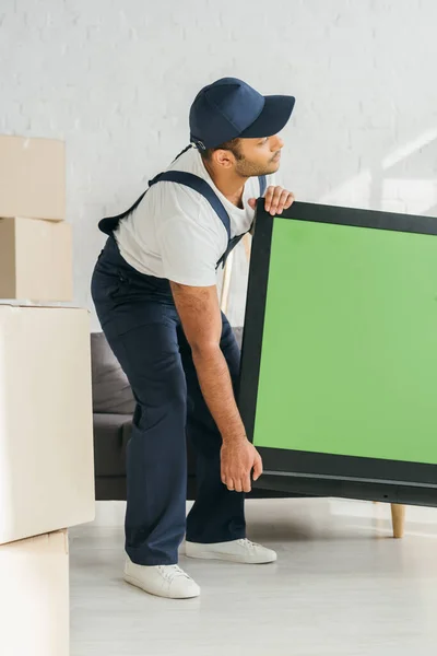 Indiano mover em uniforme transportando tv plasma com tela verde no apartamento — Fotografia de Stock