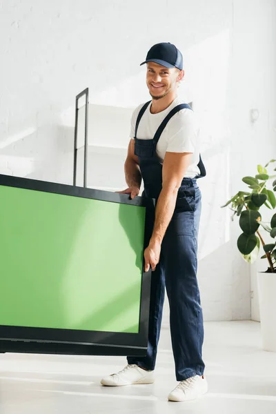 Sonriente mover en uniforme llevando plasma tv con pantalla verde en apartamento - foto de stock