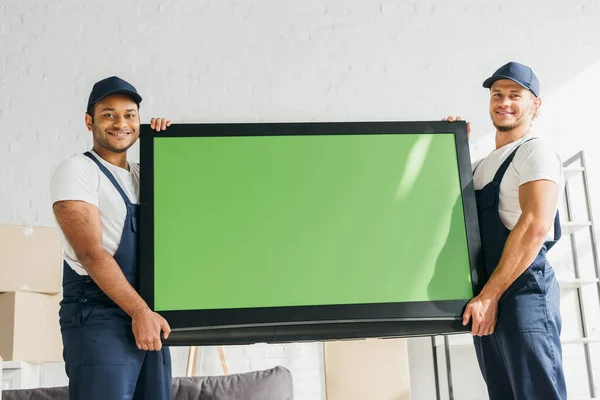 Móviles multiculturales sonrientes en uniforme llevando plasma tv con pantalla verde en apartamento - foto de stock