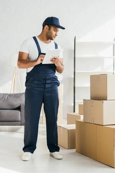 Volle Länge des indischen Arbeiters in Uniform, der ein digitales Tablet in der Hand hält und sich Boxen in der Wohnung ansieht — Stockfoto