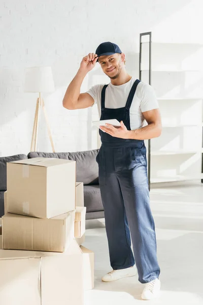 Comprimento total de movedor alegre em uniforme ajustando tampa e segurando tablet digital no apartamento — Fotografia de Stock