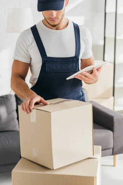 Mover in uniform holding digital tablet and looking at box in apartment — Stock Photo