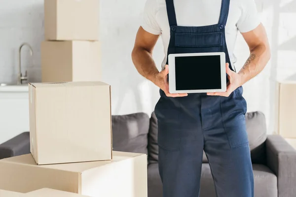 Vista recortada de la mudanza en uniforme celebración tableta digital con pantalla en blanco cerca de cajas en apartamento - foto de stock