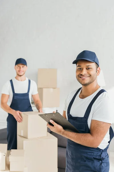 Happy indian worker holding clipboard near coworker on blurred background — Stock Photo