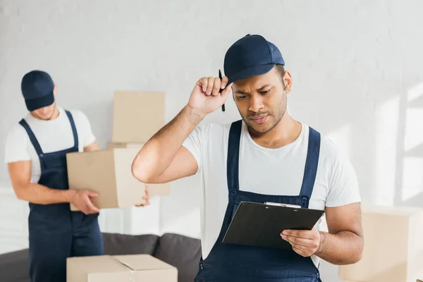 Indian worker adjusting cap while looking at clipboard near coworker on blurred background — Stock Photo