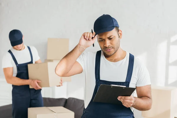 Trabajador indio ajuste tapa mientras mira portapapeles cerca de compañero de trabajo en fondo borroso — Stock Photo