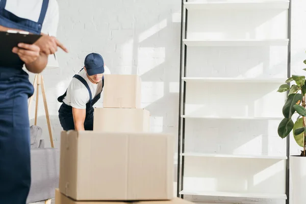 Joven transportista en tapa llevando cajas cerca de indio compañero de trabajo en fondo borroso - foto de stock