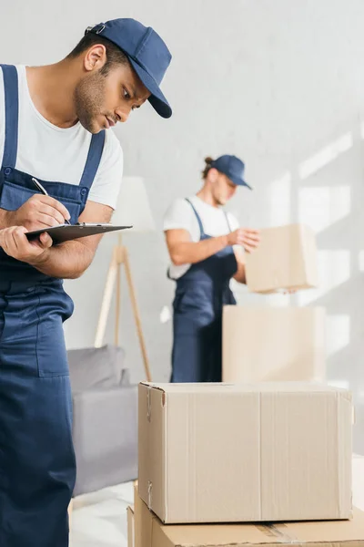 Trabajador indio sosteniendo pluma, portapapeles y mirando las cajas de cartón cerca de mover en un fondo borroso - foto de stock