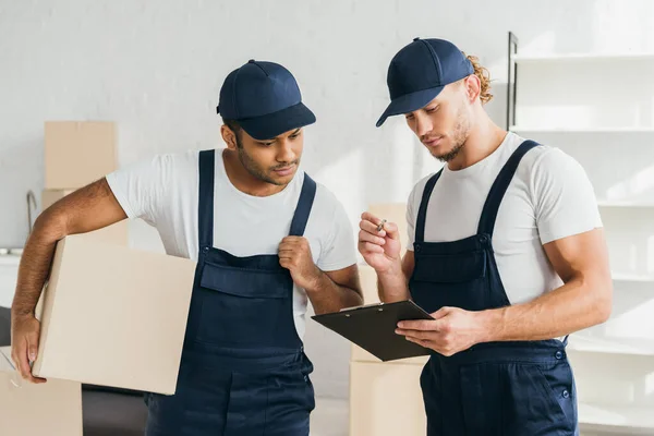 Mudanzas multiculturales mirando portapapeles en apartamento - foto de stock