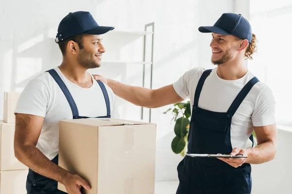 Cheerful multicultural movers smiling while looking at each other in apartment — Stock Photo