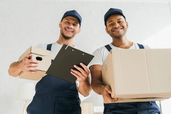 Vue à angle bas des déménageurs multiculturels joyeux souriant tout en regardant la caméra et tenant des boîtes dans l'appartement — Photo de stock