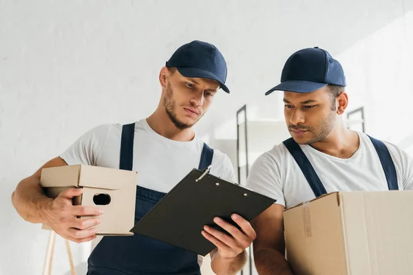 Traslocatori multiculturali che tengono scatole e guardando appunti in appartamento — Foto stock