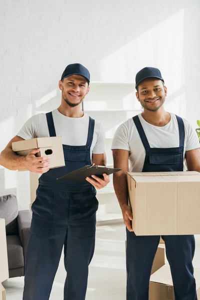 Cheerful multicultural movers smiling while looking at camera and holding boxes in apartment — Stock Photo