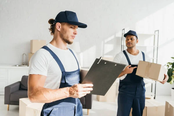 Man in cap and uniform looking at clipboard near indian coworker pointing with finger at box on blurred background — Stock Photo