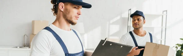 Hombre de gorra y uniforme mirando portapapeles cerca de indio compañero de trabajo señalando con el dedo en la caja sobre fondo borroso, pancarta — Stock Photo