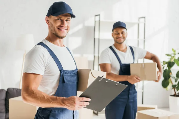 Hombre feliz en gorra y uniforme sujetando portapapeles cerca de compañero de trabajo indio caja de espera sobre fondo borroso - foto de stock