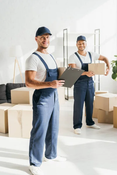 Longitud completa del hombre feliz en la tapa y el portapapeles de sujeción uniforme cerca de compañero de trabajo indio que sostiene la caja en fondo borroso - foto de stock